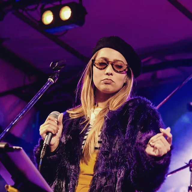 ROMII on stage in a purple faux fur coat and beret, holding a microphone